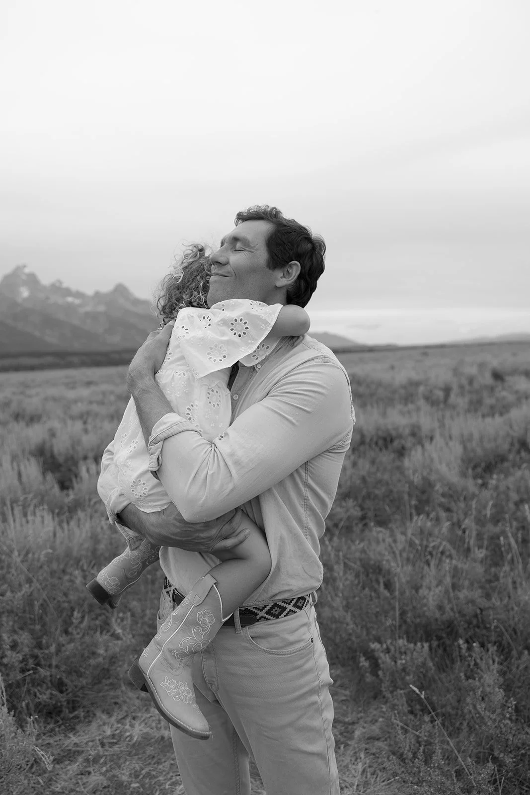 A man holding a young girl in a field, embracing and smiling with eyes closed, mountain range in the background, in black and white.