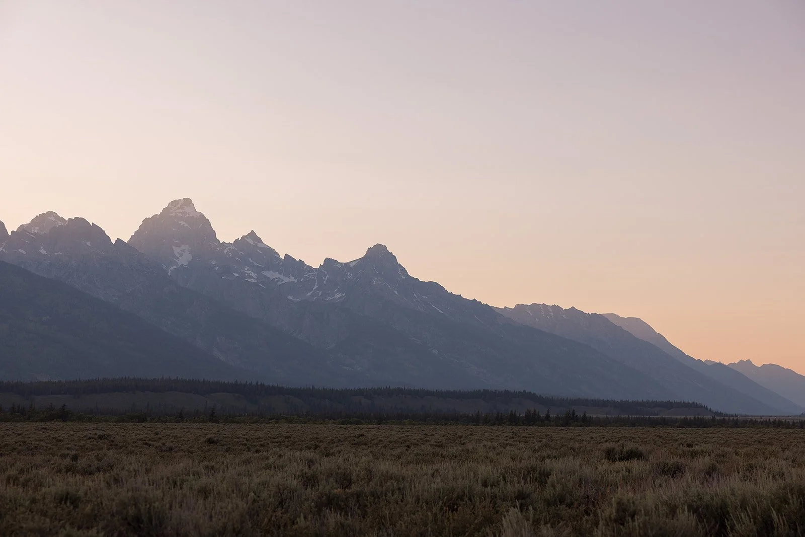 Mountain range at dusk with snow-capped peaks and a flat, grassy plain in the foreground.