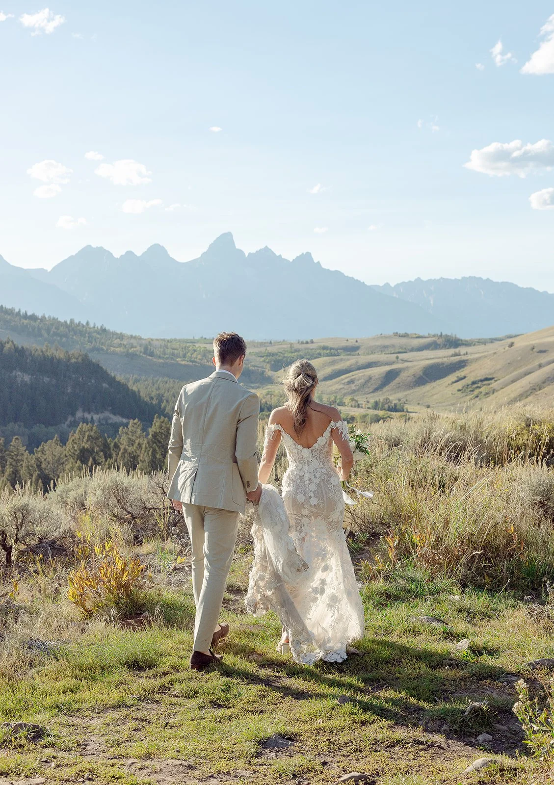 A bride and groom walking hand in hand in a scenic outdoor location with mountains in the background on their wedding day.