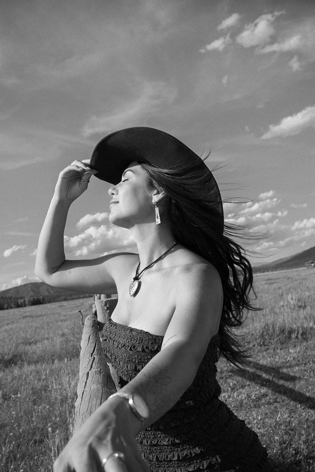 Woman in a black dress and wide-brimmed hat standing outdoors with her eyes closed, holding her hat, and taking a selfie in a grassy field with mountains and a cloudy sky in the background.