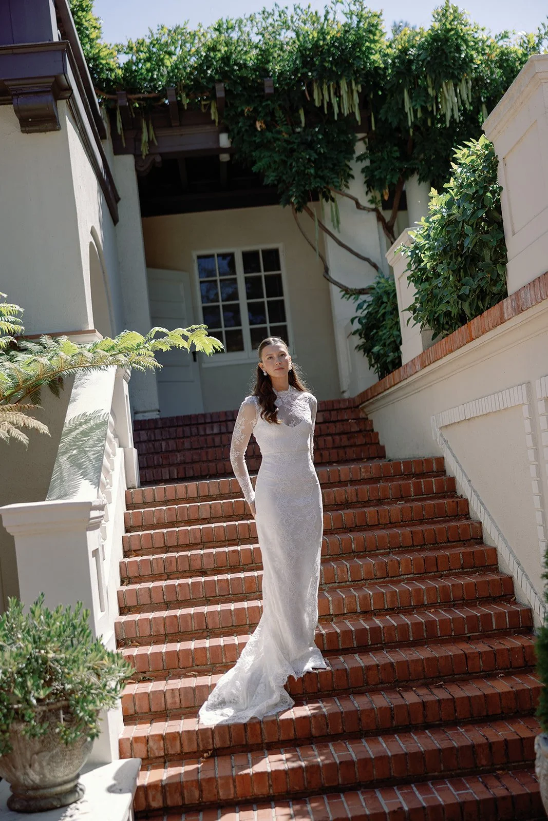 Woman in white lace dress standing on brick staircase with greenery and building in the background.