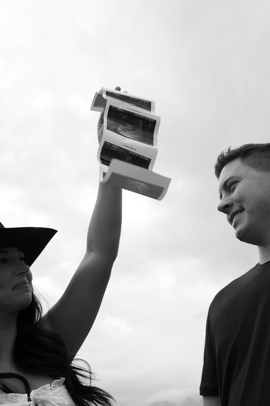 A woman holding up printed pages or posters with her right hand, while a man looks at her and smiles. The woman wears a hat, and the background consists of a cloudy sky.