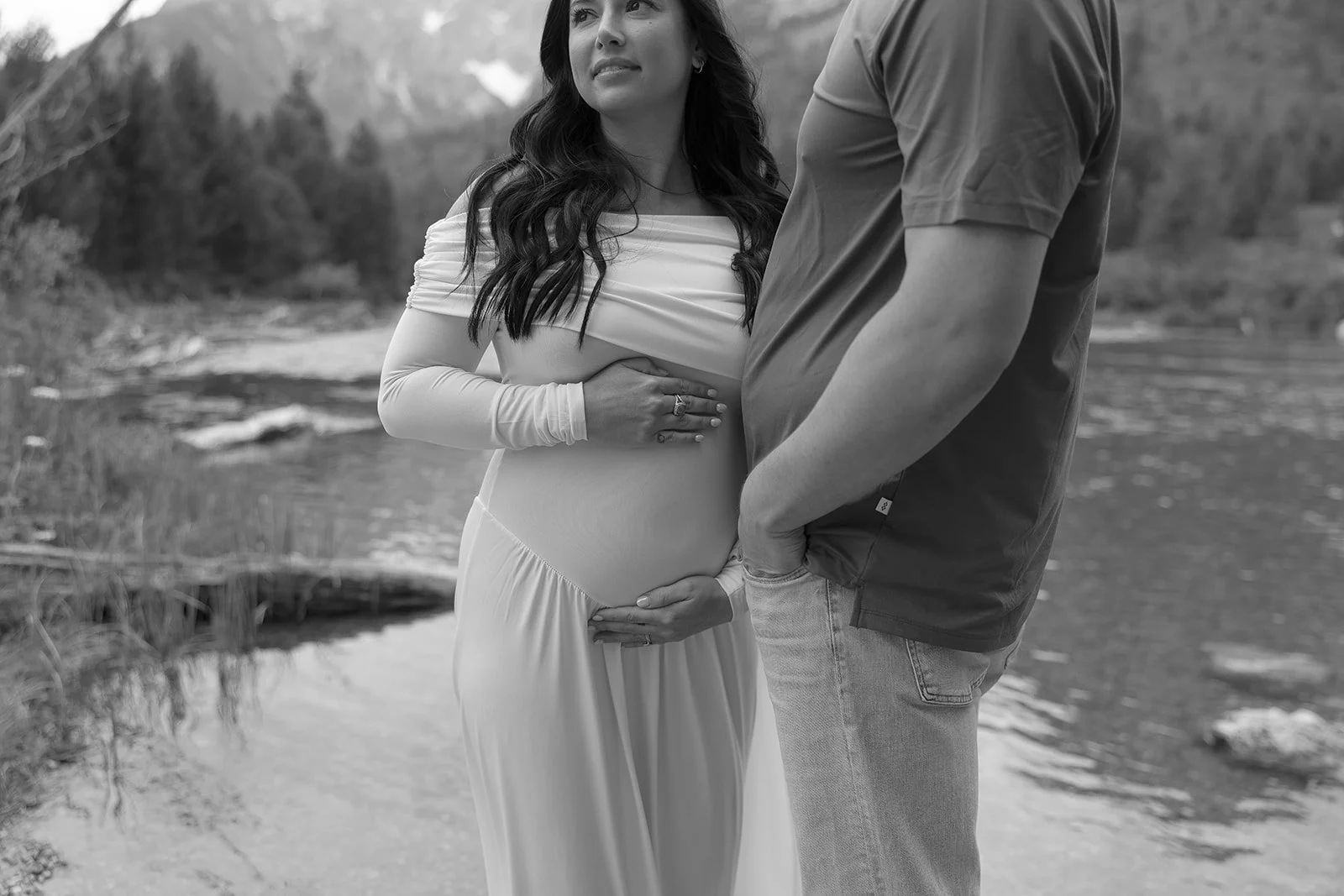 A pregnant woman in a long dress stands outdoors near a body of water with a man, both holding her belly and chest.