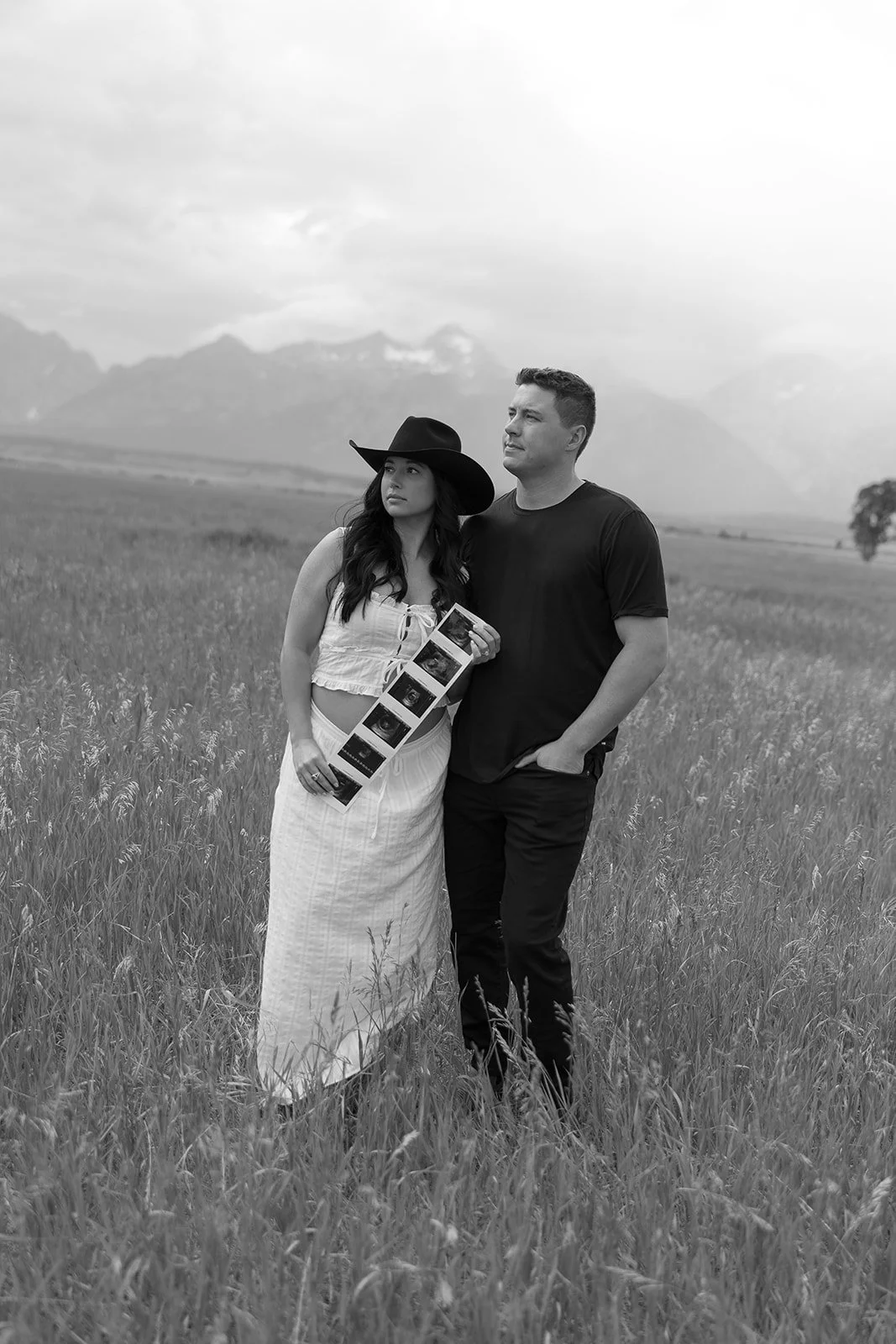 A woman and man standing in a grassy field, woman holding a strip of photographs, woman wearing a wide-brimmed hat, and mountain range in the background.