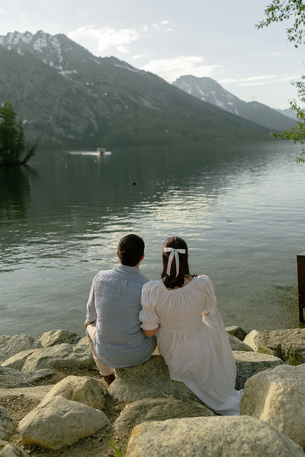 A couple sitting on rocks by a lake, facing mountains, during daylight.