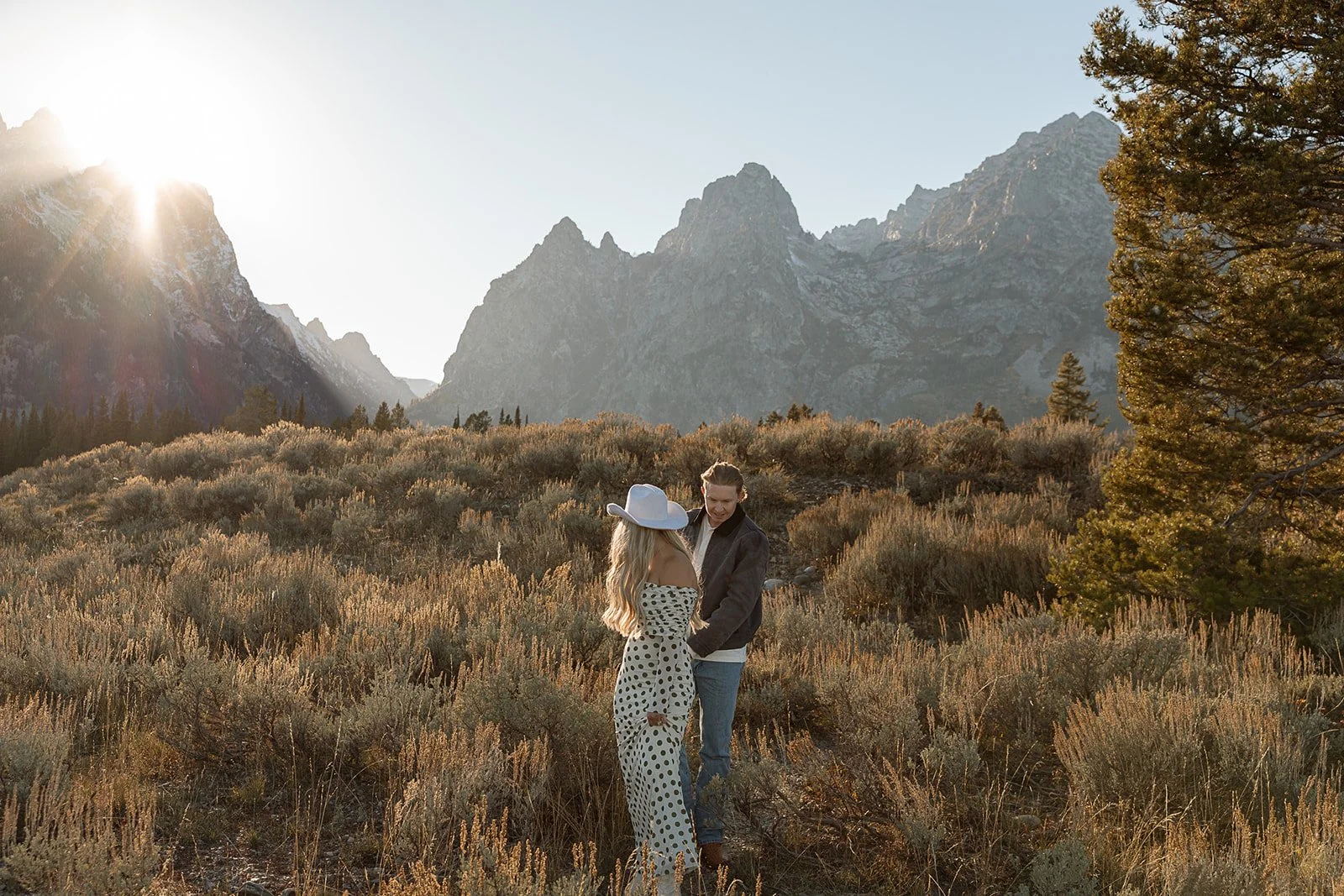 A couple standing in a field of dry bushes with mountains and a clear sky in the background, the sun shining from the left side.