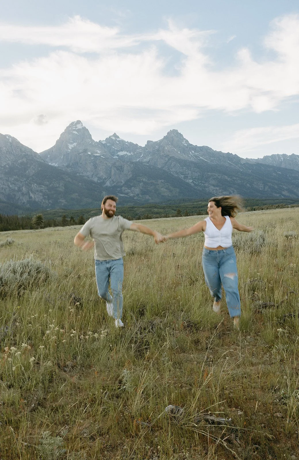 A happy couple holding hands and running through a grassy field with mountains in the background.