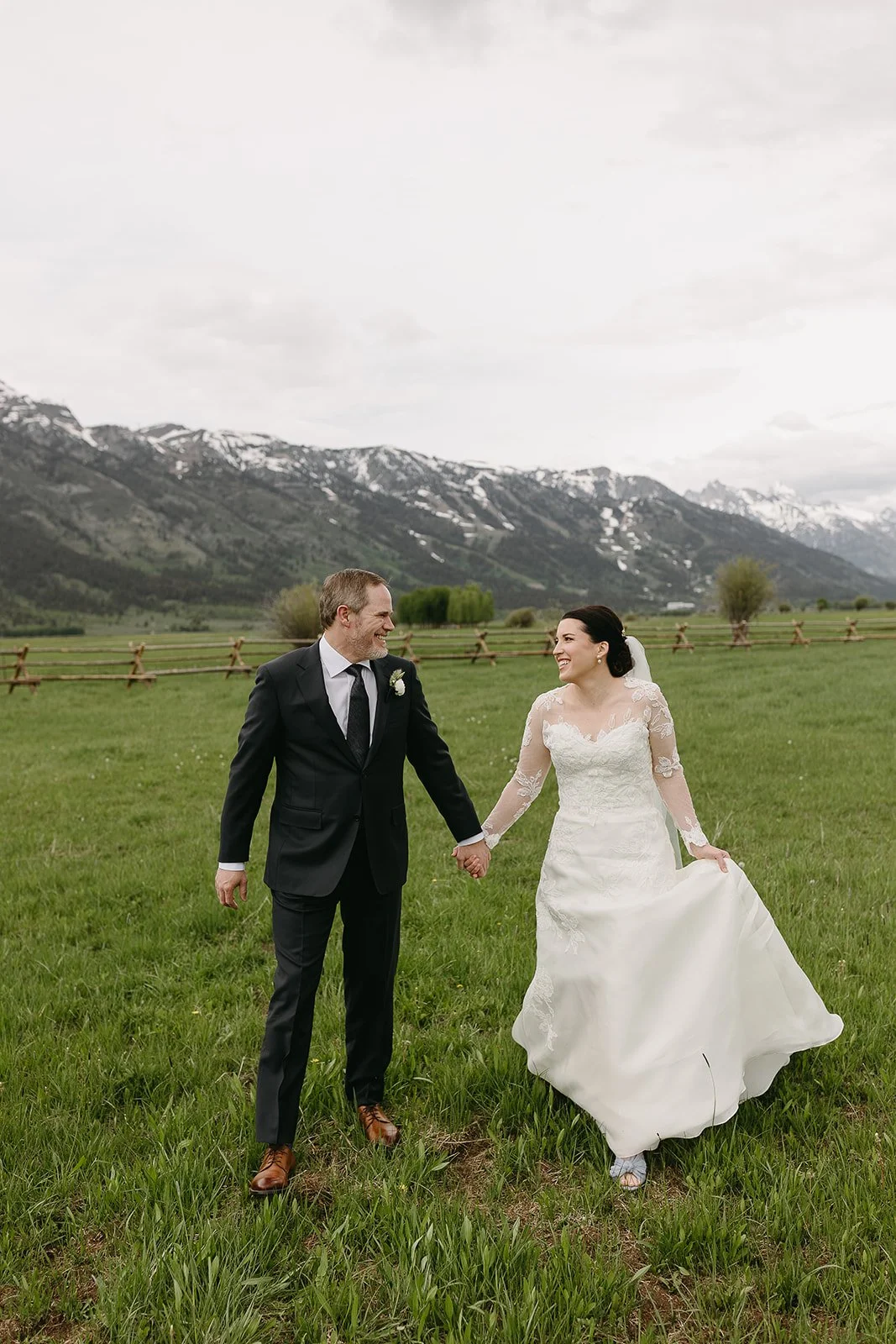 A bride and groom holding hands and smiling at each other in a green field with mountains in the background.
