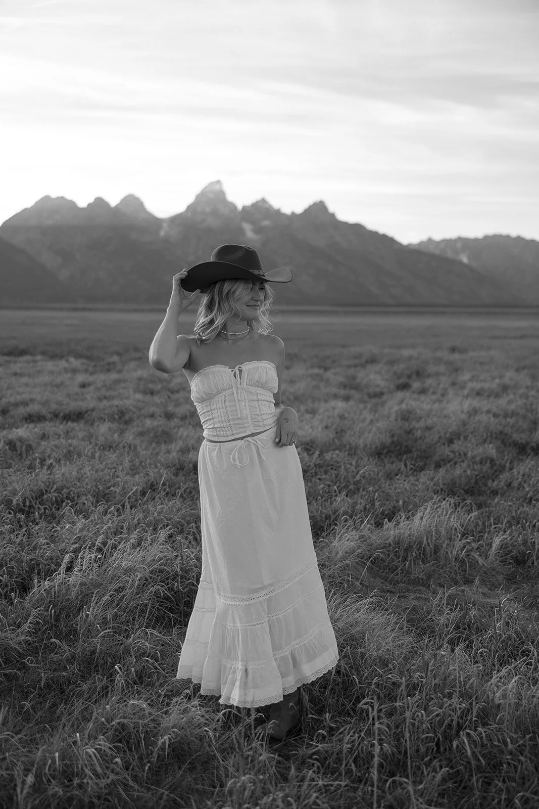 A woman standing in a grassy field holding a cowboy hat, with mountains in the background, photographed in black and white.