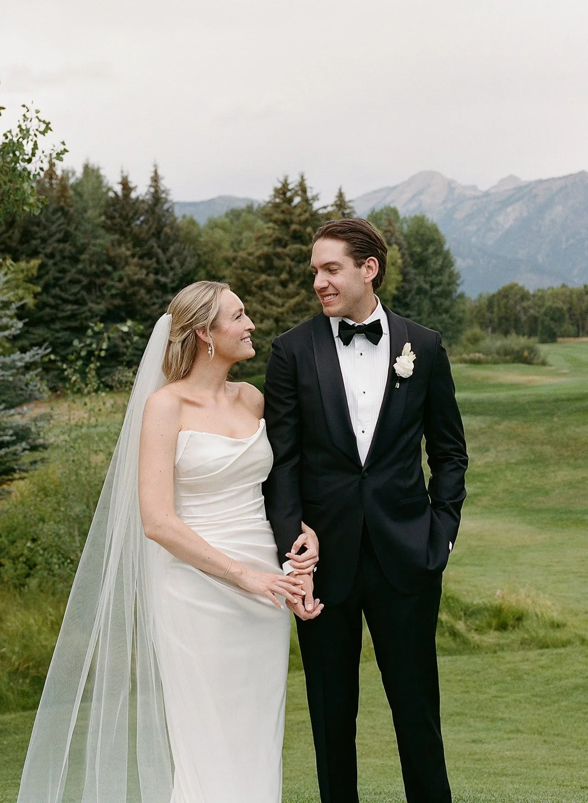 A bride and groom standing outdoors on a golf course, holding hands and smiling at each other, with mountains and trees in the background.