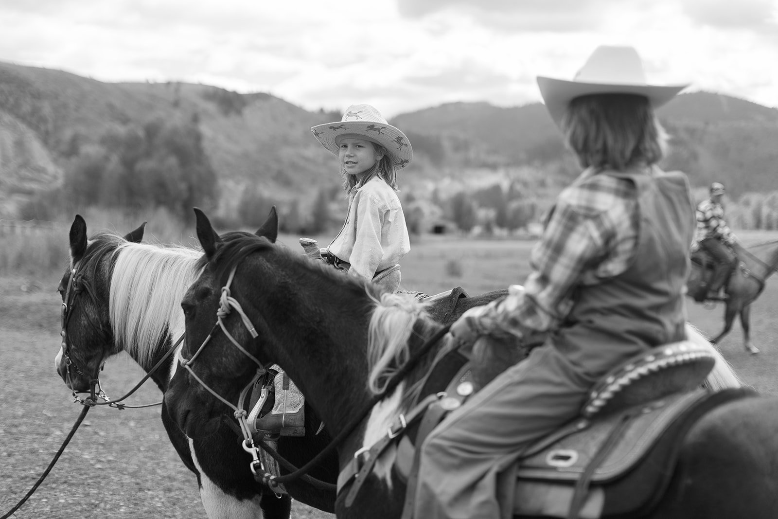A group of children riding horses outdoors, wearing cowboy hats, with a mountainous landscape in the background.
