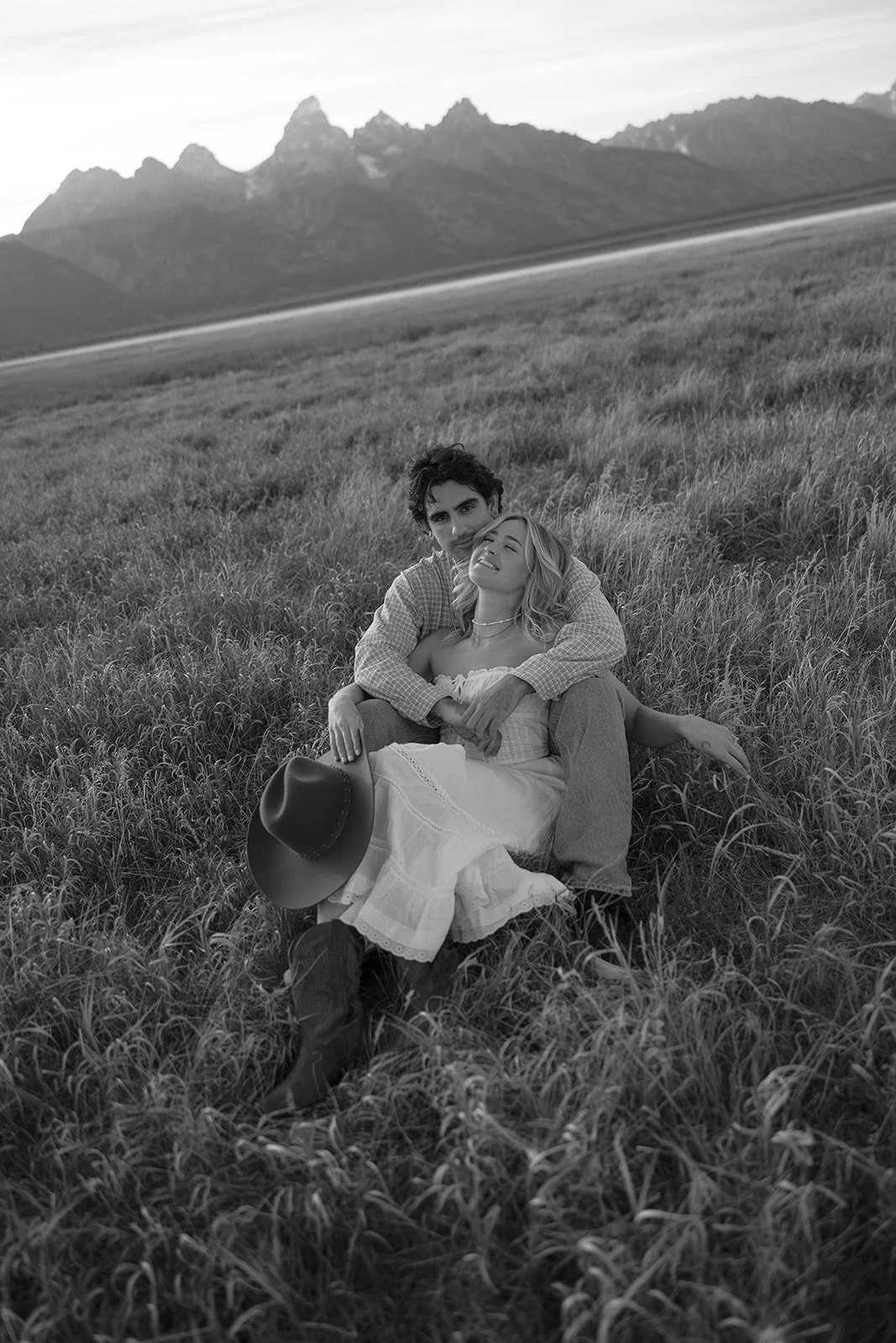 A black and white photo of a couple sitting in tall grass in a field, with mountain peaks in the background. The woman is smiling with eyes closed, holding a hat, while the man is behind her, embracing her.