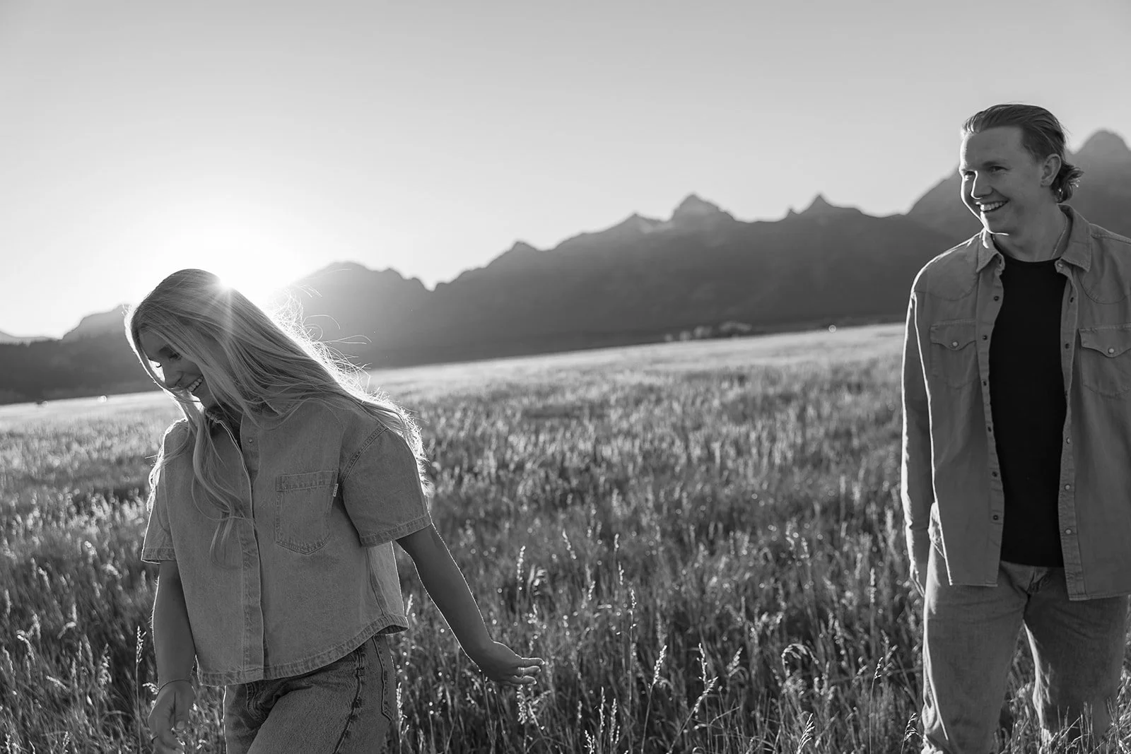A black and white photo of a woman and a man walking in a field of tall grass with mountains in the background, sunlight shining from behind the mountains.