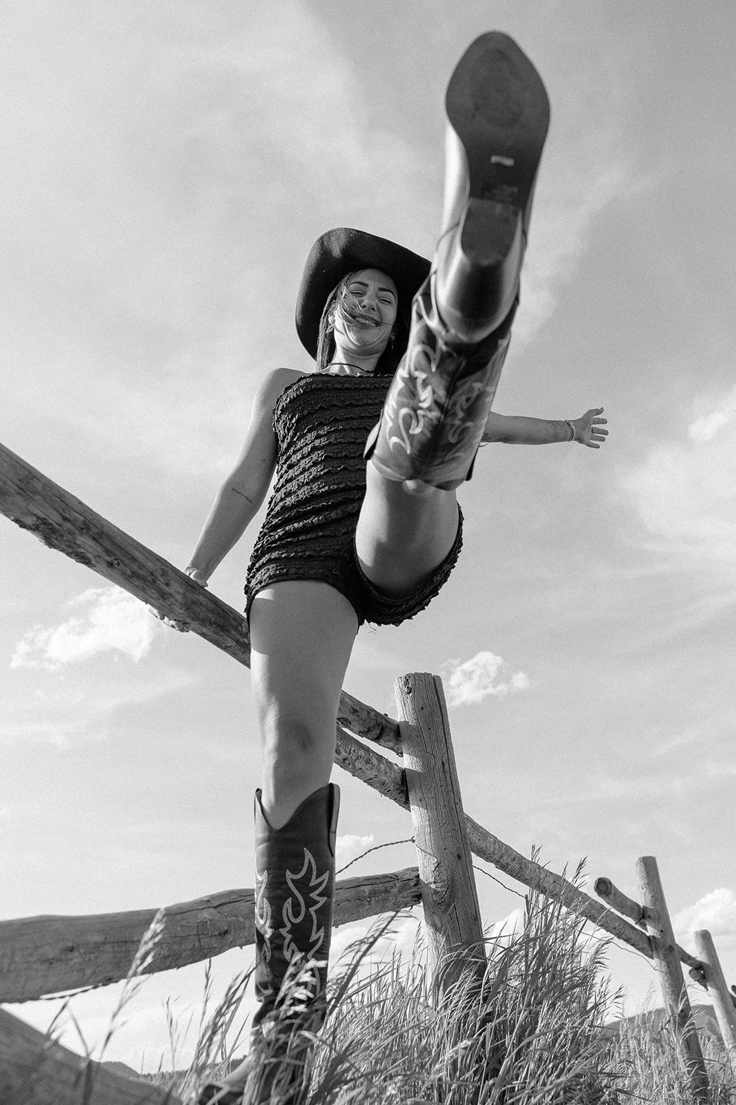 A woman wearing a hat, black strapless top, and cowboy boots, standing on a wooden fence post, lifting her leg high, outdoors with a cloudy sky and grass in the background.