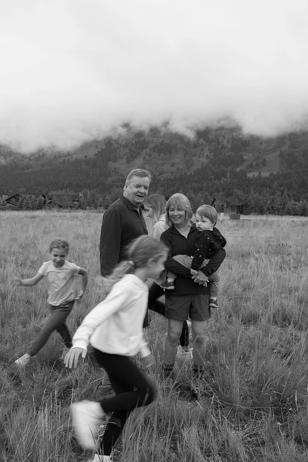 A black and white photo of a family of six outdoors in a grassy field with mountains and clouds in the background. Two adults, three children, and a toddler are present. The woman is holding the toddler, and the young girl appears to be running. The 