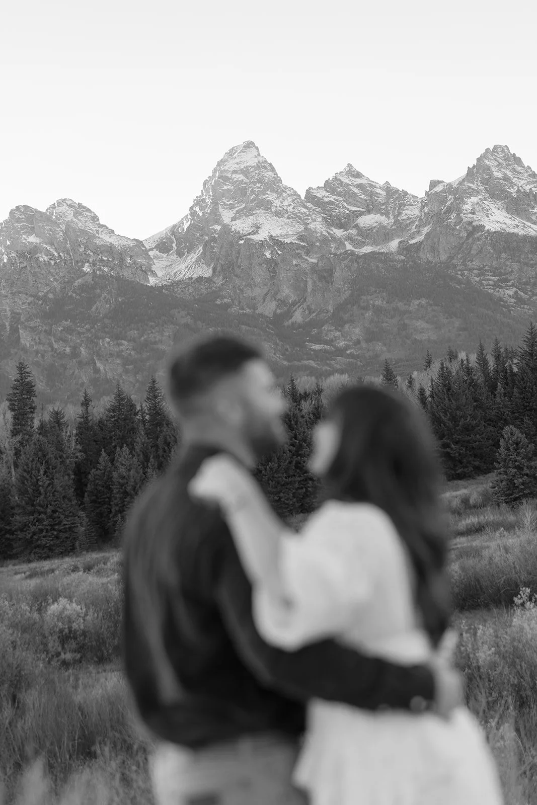 A black-and-white photograph of a couple in the foreground, blurry, facing each other, with a mountain range and pine forest in the background.