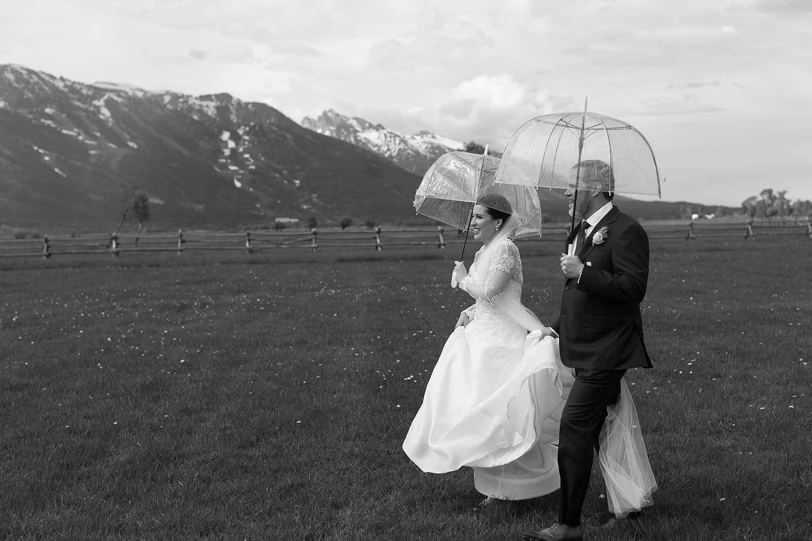 A bride and groom walking outdoors while holding umbrellas on a grassy field, with mountains in the background, in black and white.