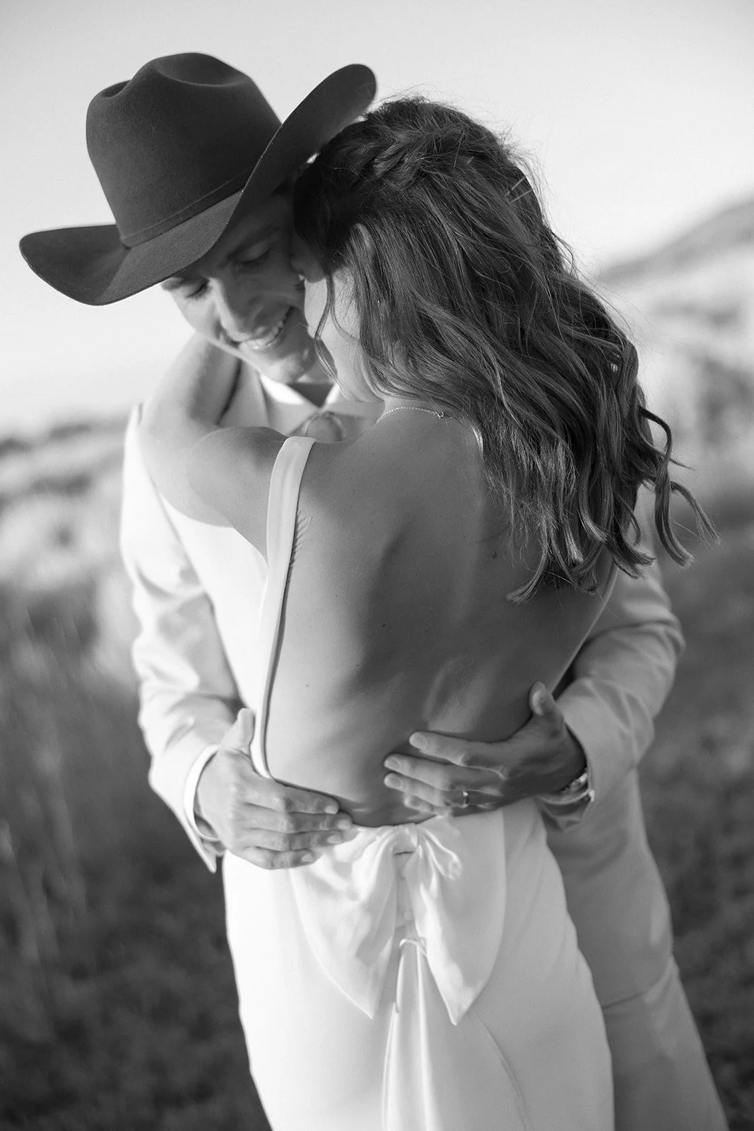 A couple embraces outdoors, with the man wearing a cowboy hat and the woman dressed in a backless dress, sharing a tender moment.