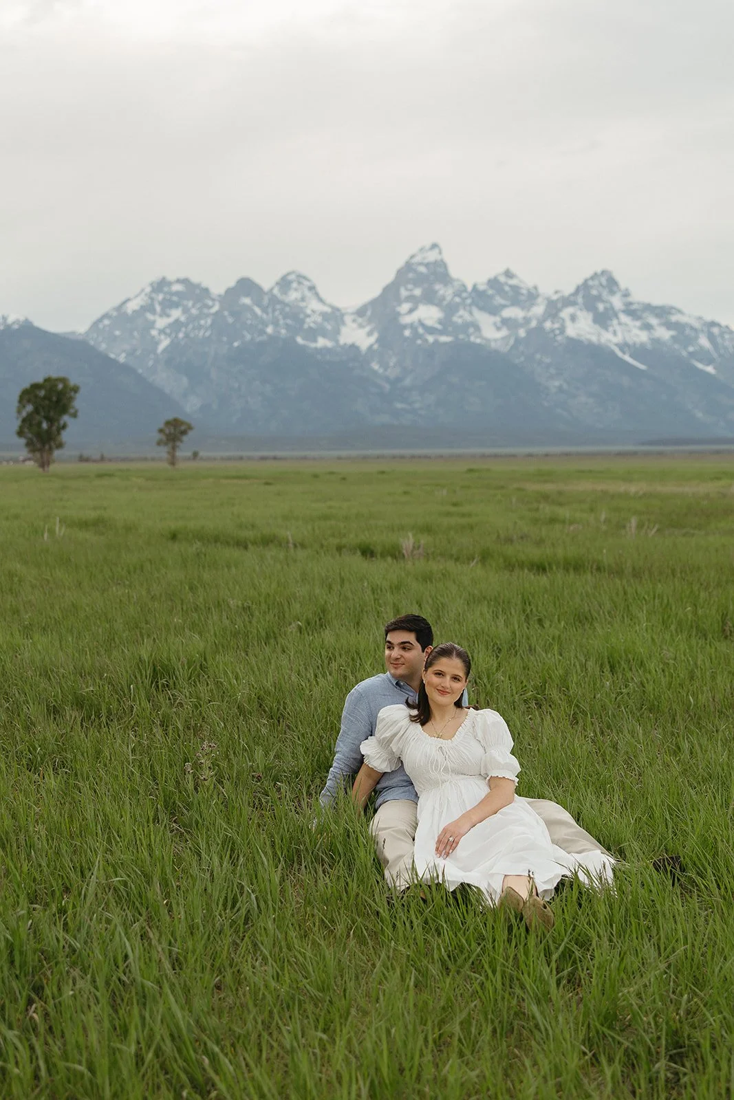 A couple sitting in tall green grass in a field, with a backdrop of distant mountains and overcast sky.
