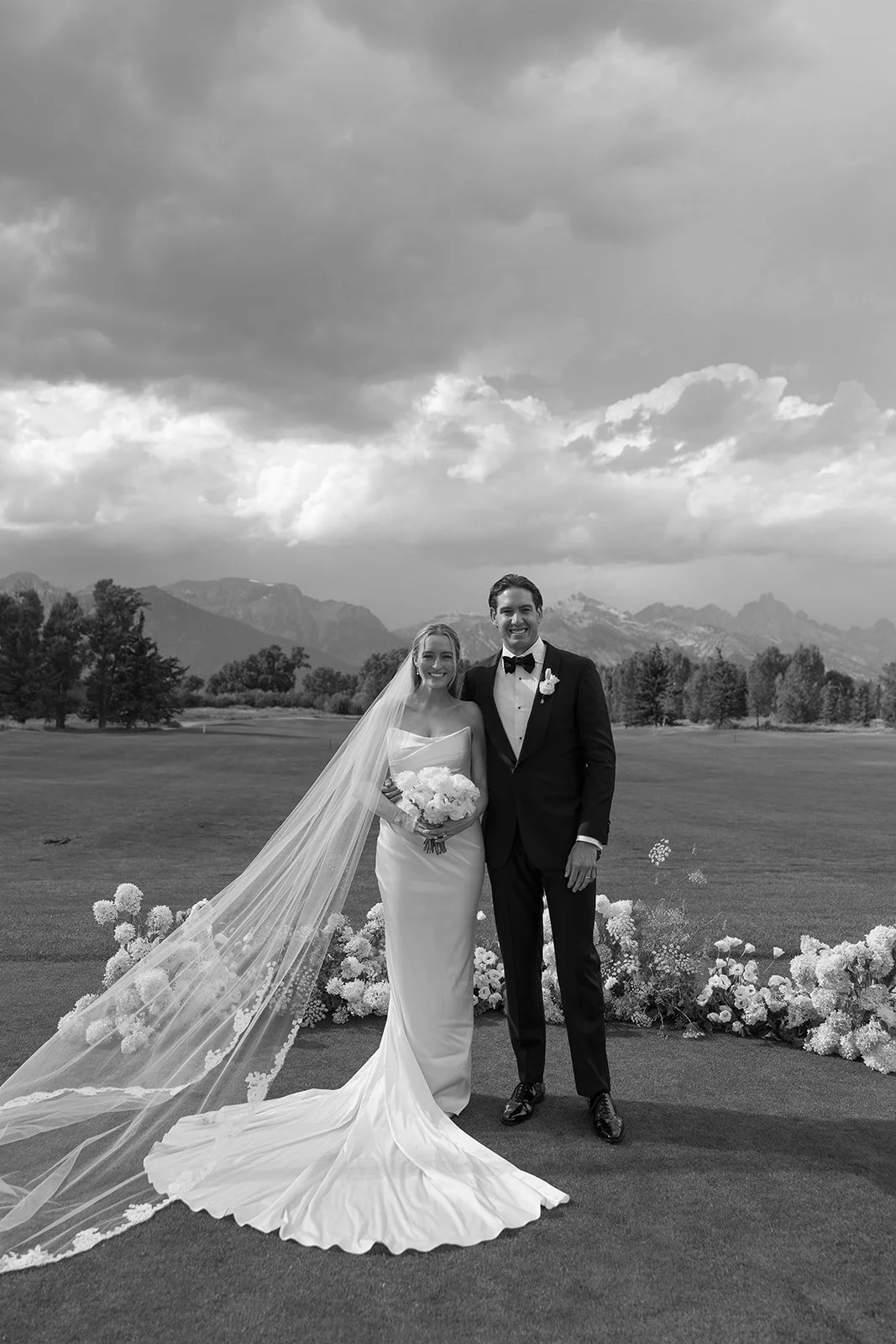 Black and white photo of a bride and groom standing outdoors on a wedding day, with mountains and cloudy sky in the background. The bride holds a bouquet, and the groom is dressed in a tuxedo with a bow tie.