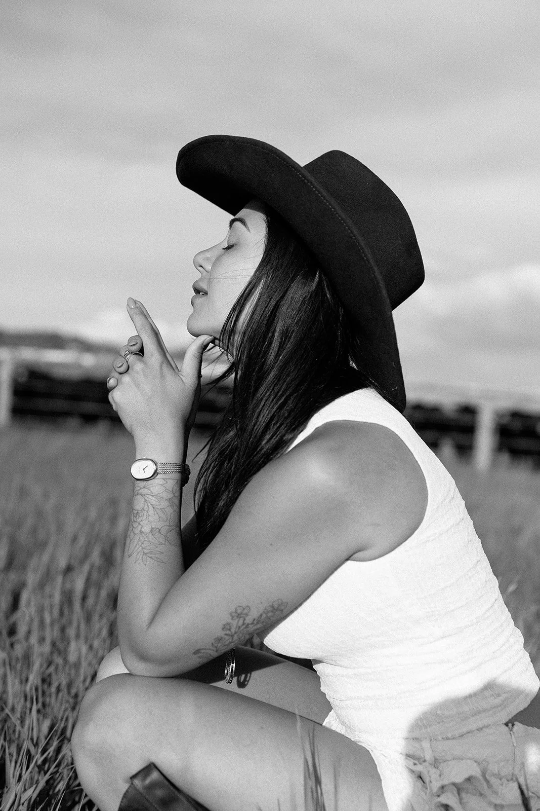 A woman wearing a wide-brimmed hat and sleeveless top sitting in a field of tall grass, with her eyes closed and hands held near her face, captured in black and white.