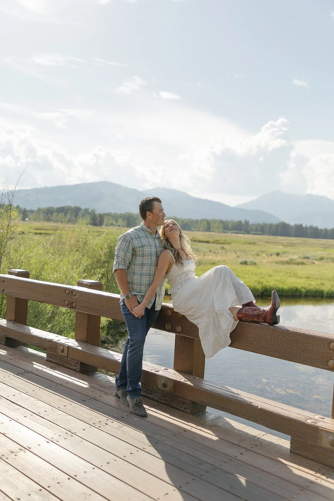 A man and woman standing and sitting on a wooden bridge overlooking a river, with a scenic landscape of green fields and mountains in the background. The woman is wearing a white dress and cowboy boots, leaning back with her head tilted, while the ma