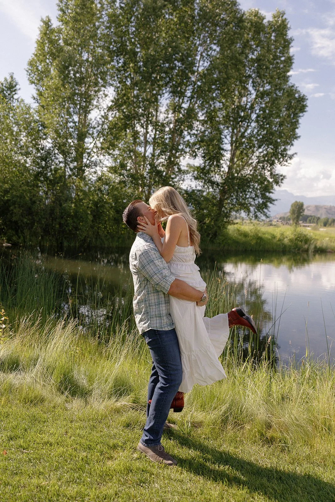 A man lifting a woman in a white dress near a lake with trees in the background on a clear day.