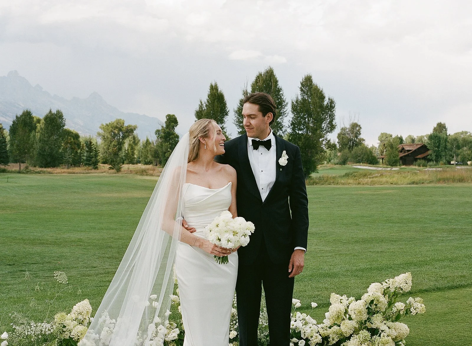 A bride and groom standing close together outdoors on a wedding day, with greenery and trees in the background, the bride holding a bouquet of white flowers, both smiling at each other.