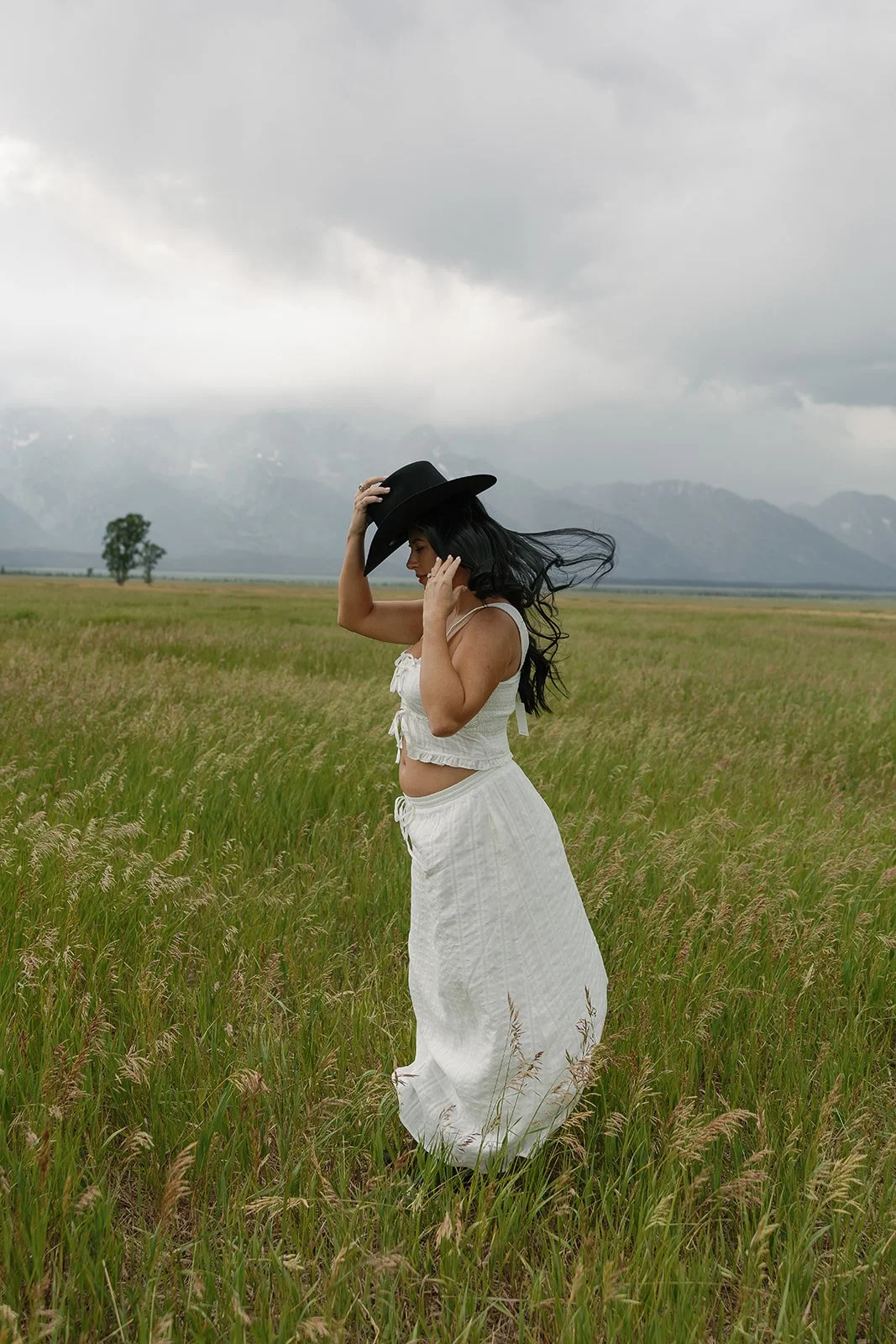 A woman in a white outfit and black hat walking through a grassy field on a cloudy day, with mountains in the background.