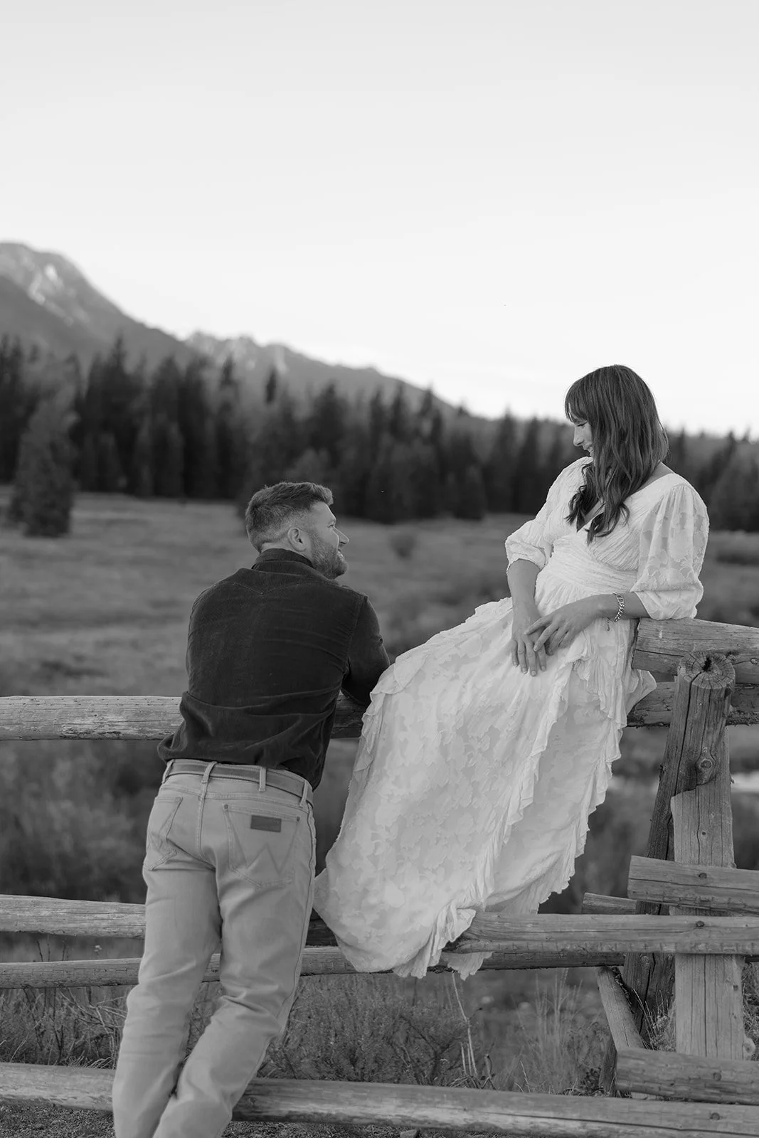 A man and pregnant woman in a scenic outdoor setting, with mountains and trees in the background. The woman sits on a wooden fence in a white dress, while the man stands close, facing her and smiling.