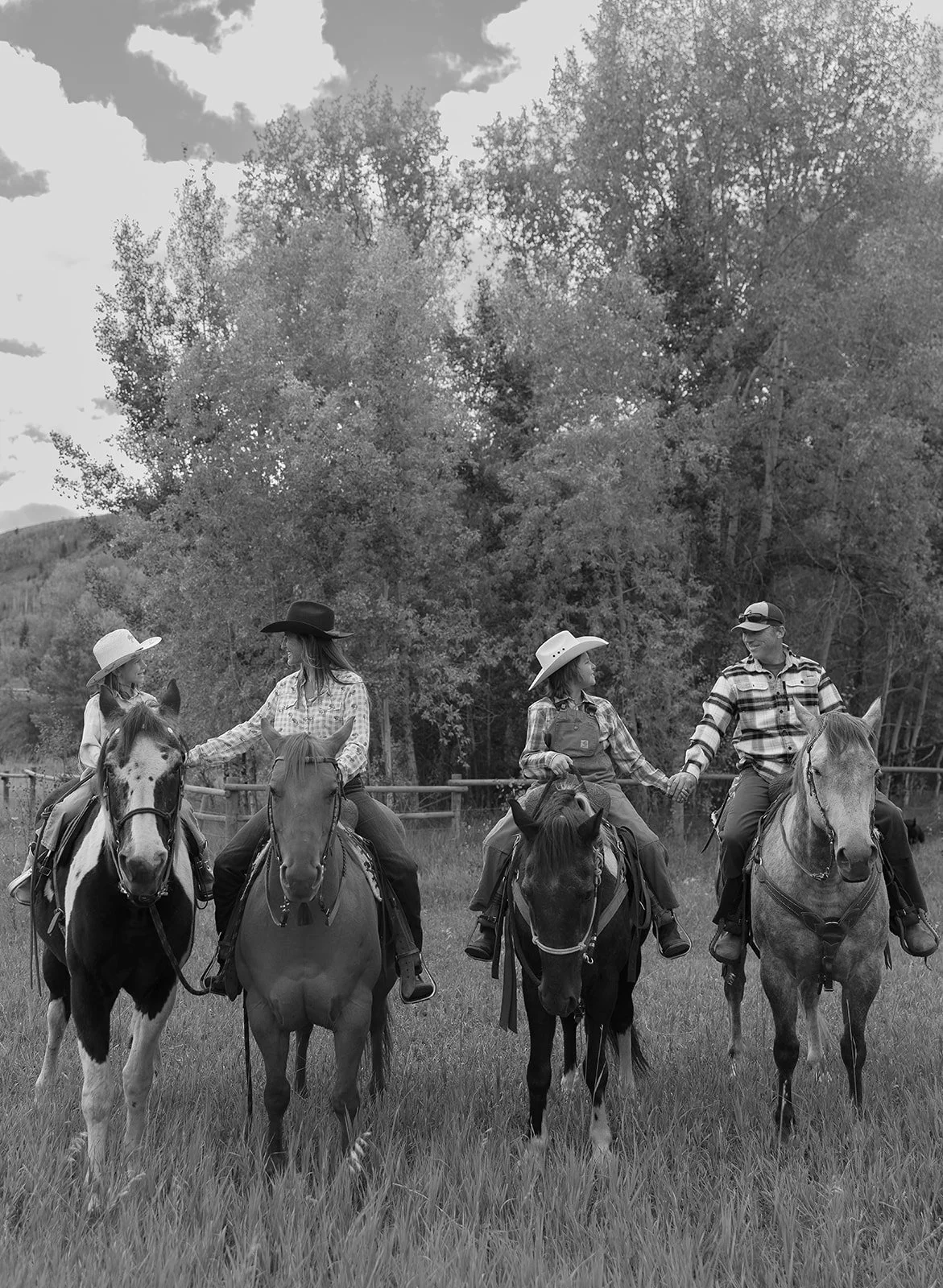 Four people riding horses in a grassy area, with two women wearing cowboy hats and a man wearing a baseball cap, holding hands and interacting.