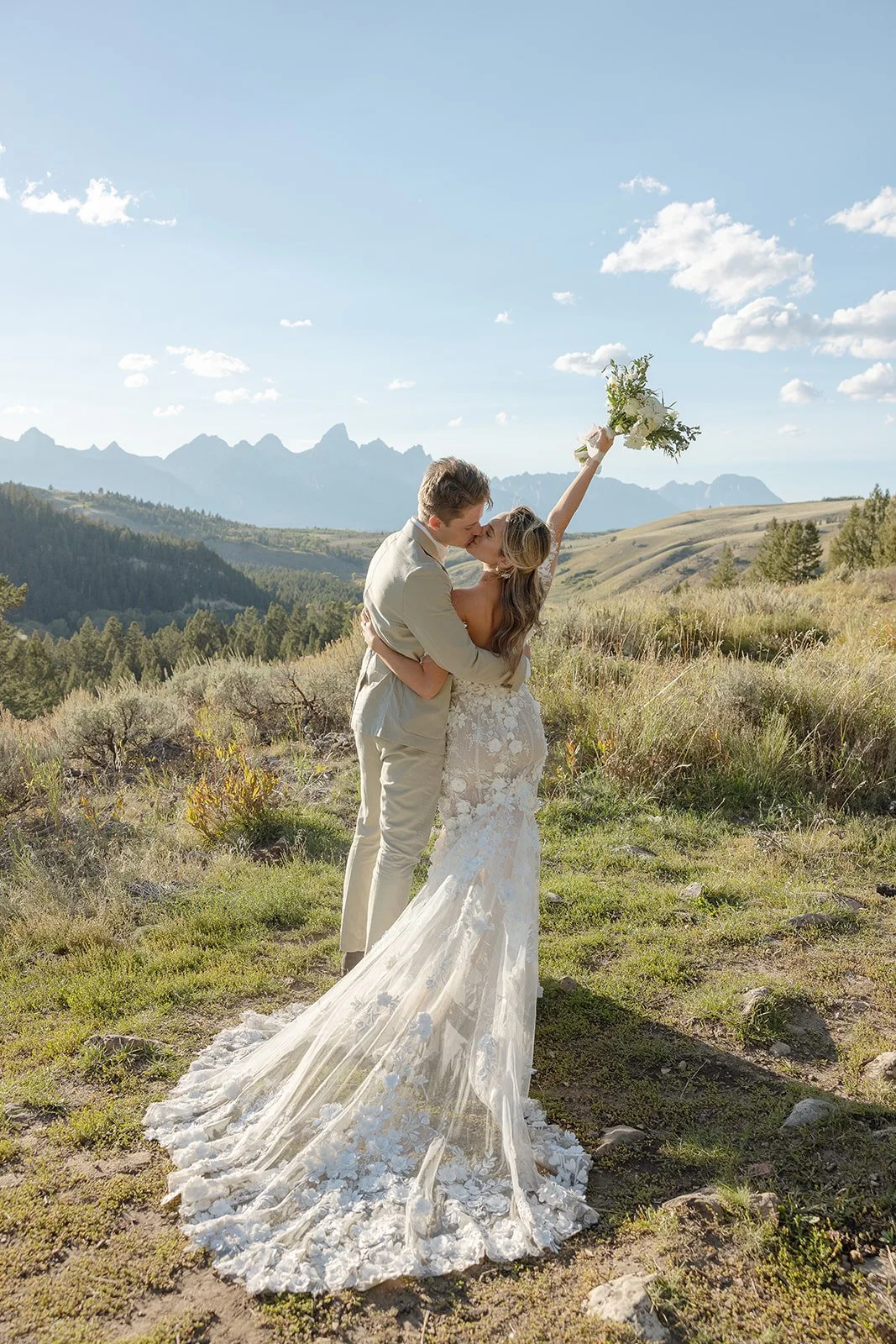 A couple in wedding attire sharing a kiss in an outdoor scenic landscape with mountains and hills in the background. The bride is holding a bouquet and has her arm raised above them.