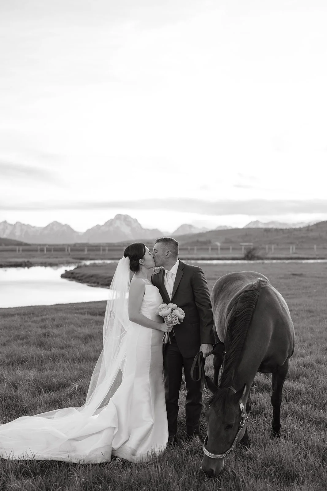 A black and white photo of a bride and groom kissing outdoors near a horse, with mountains and a lake in the background.