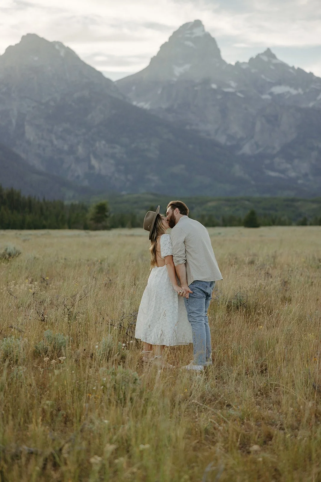 A couple kissing in a grassy field with mountains in the background.