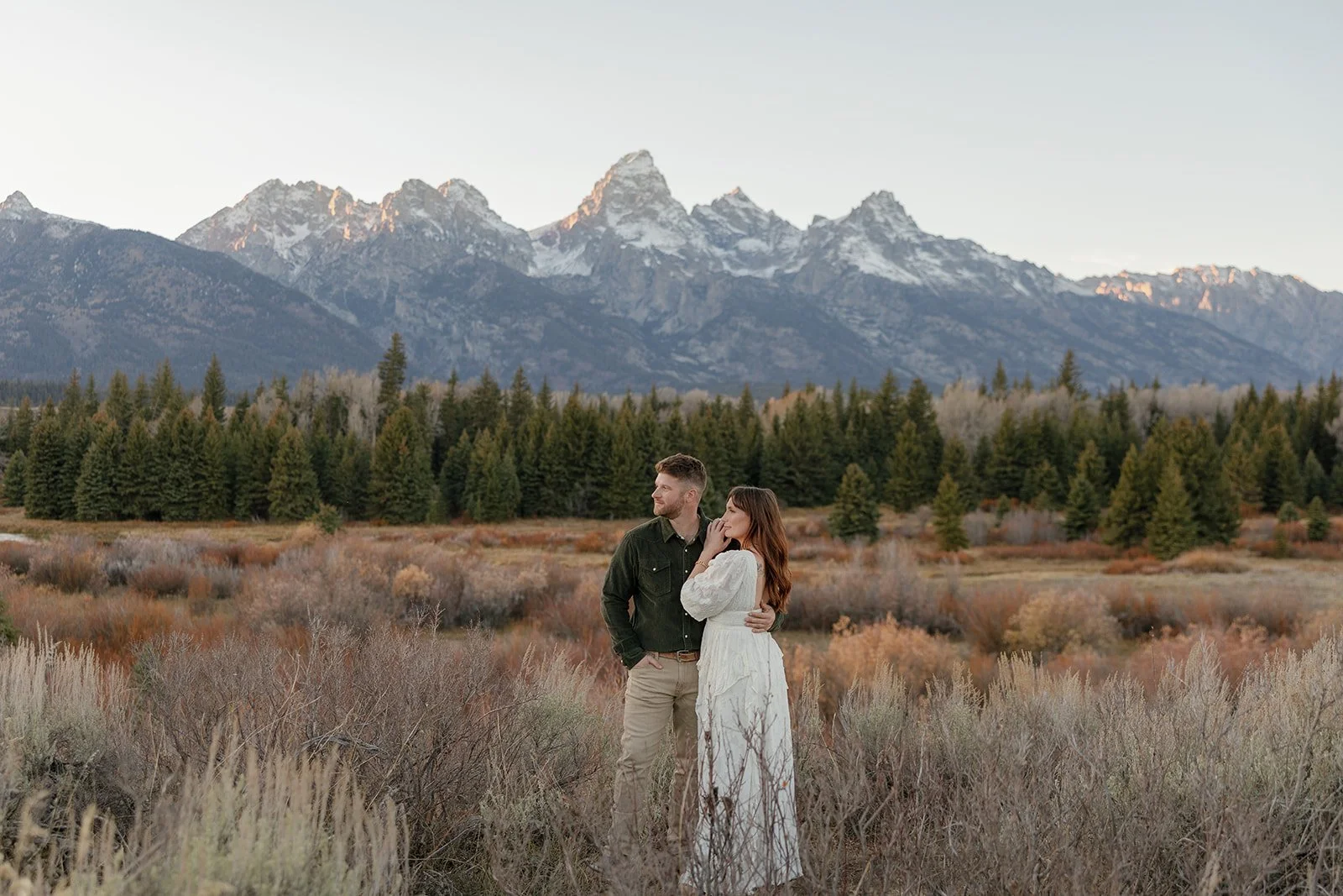 A couple standing in a field with mountains and trees in the background during sunset or early evening.