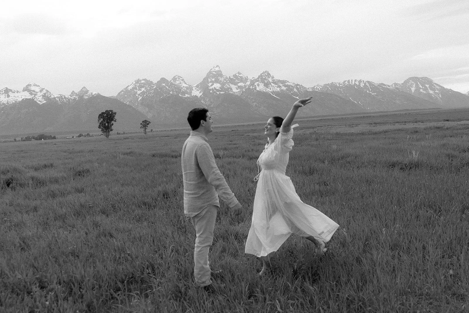 A couple holding hands in a grassy field with mountains and snow-capped peaks in the background