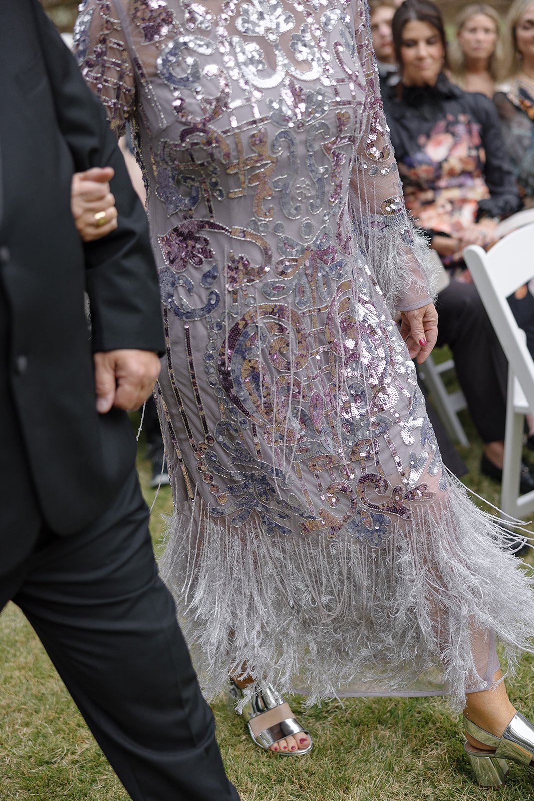 Woman in a sequined, fringed dress walking next to a man in formal attire, with seated spectators in the background.