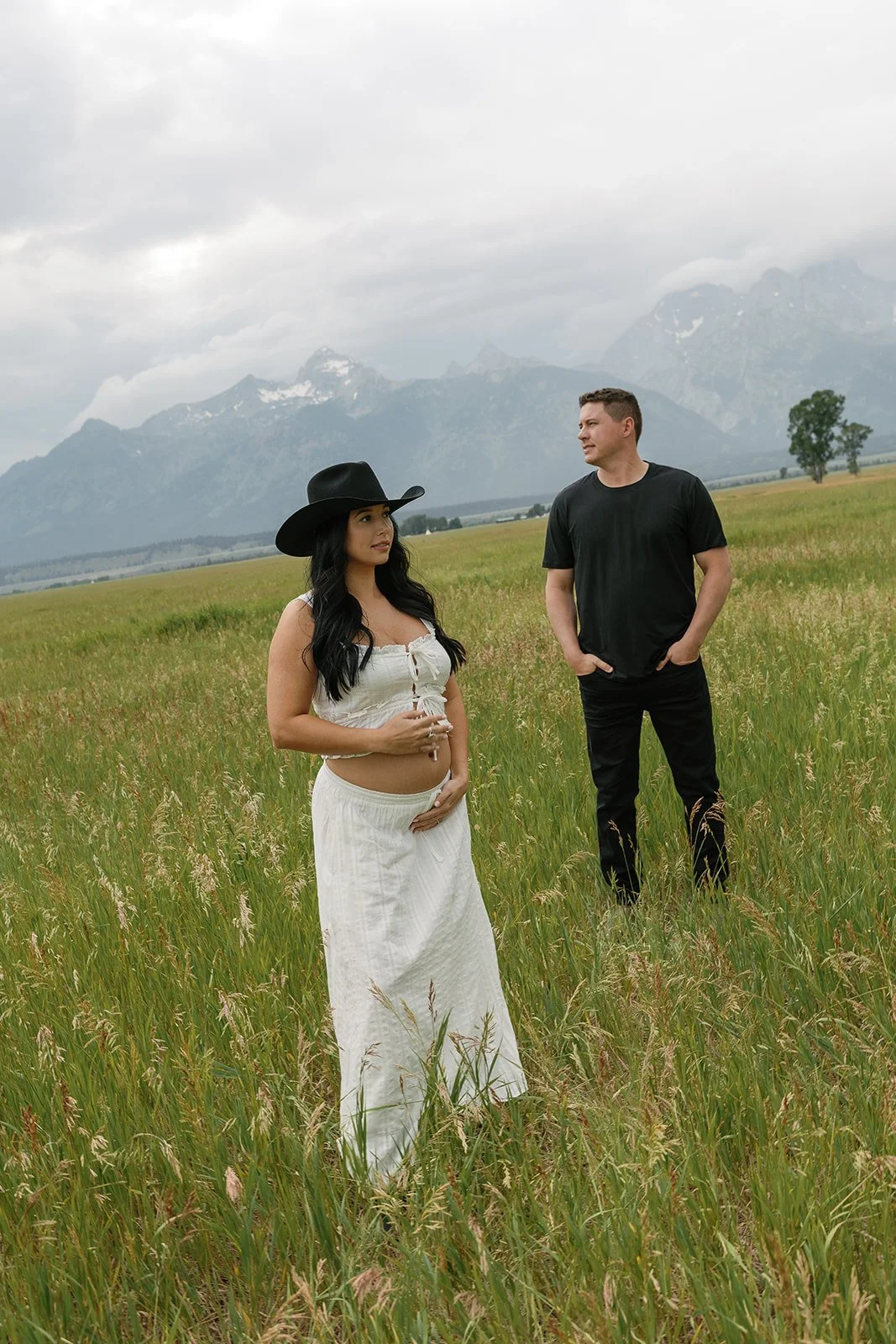 A woman in a white skirt and black cowboy hat stands in a green field with tall grass, while a man in a black t-shirt and black pants stands nearby. Mountains are visible in the background, and the sky is cloudy.