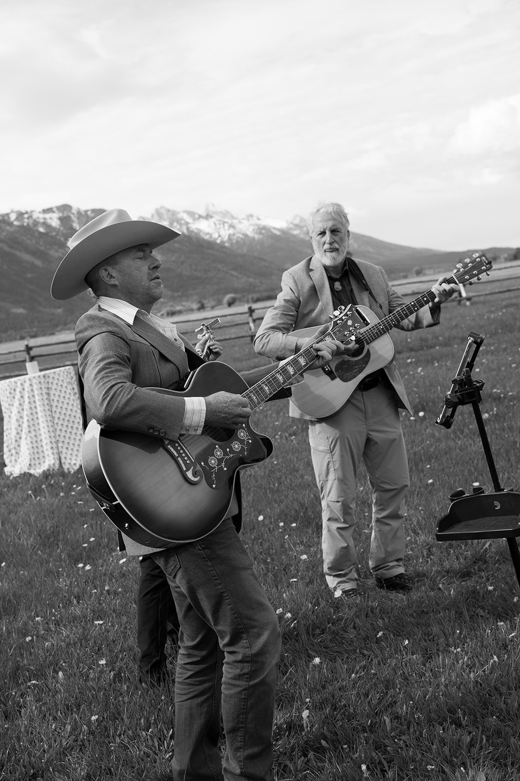 Two men playing guitars outdoors in a grassy field with mountains in the background, one wearing a cowboy hat.