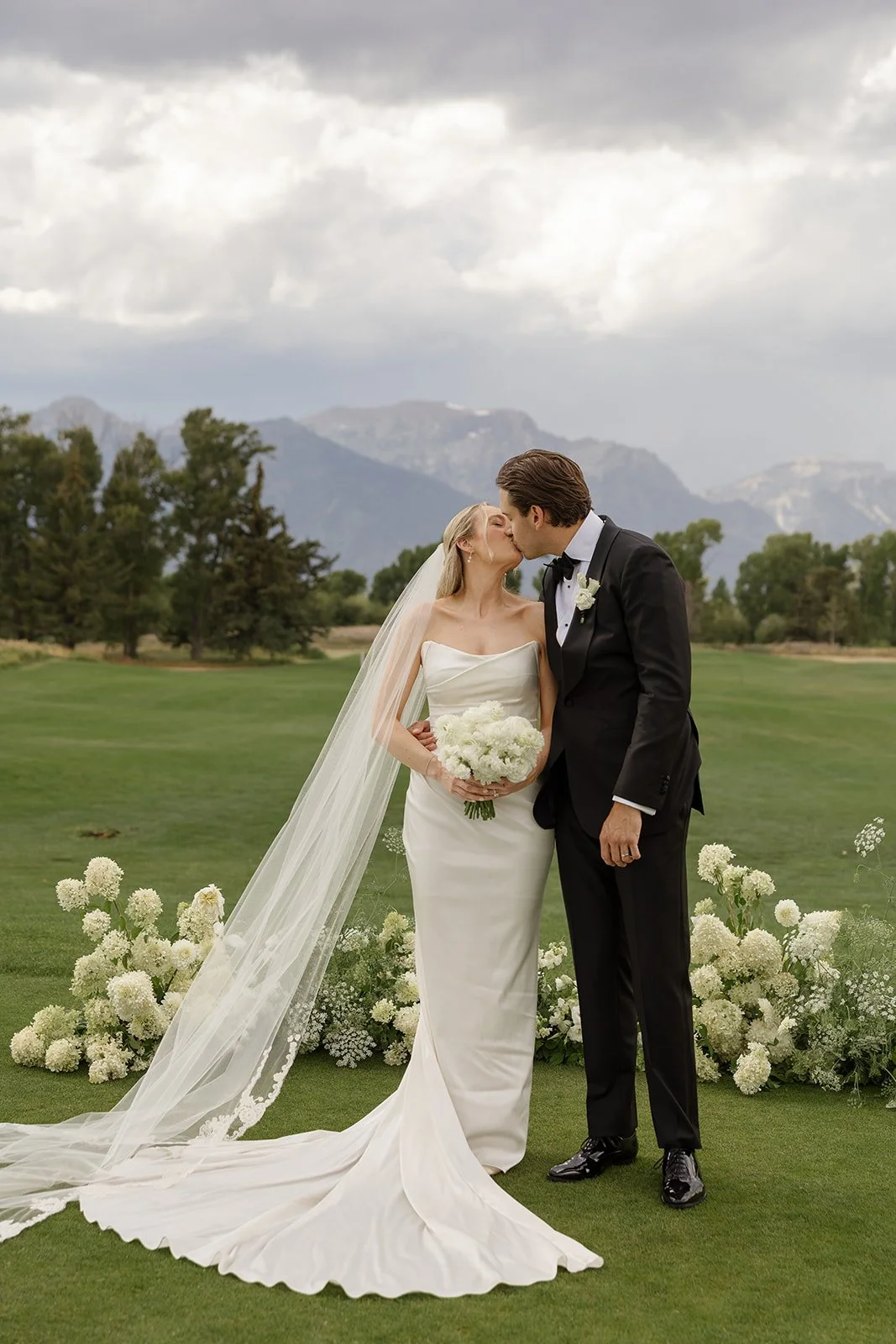 A bride and groom sharing a kiss at their wedding outdoors, with mountains and trees in the background, flowers on the ground, and cloudy sky overhead.