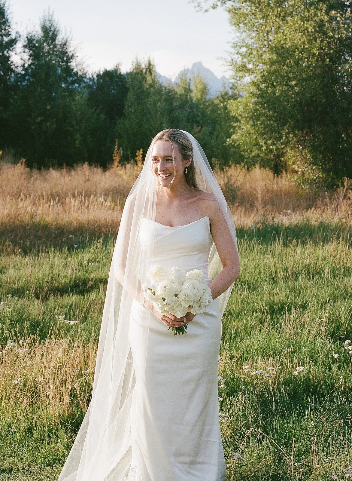 A smiling bride in a white wedding dress and veil holding a bouquet of white flowers in a grassy field with trees and mountains in the background.