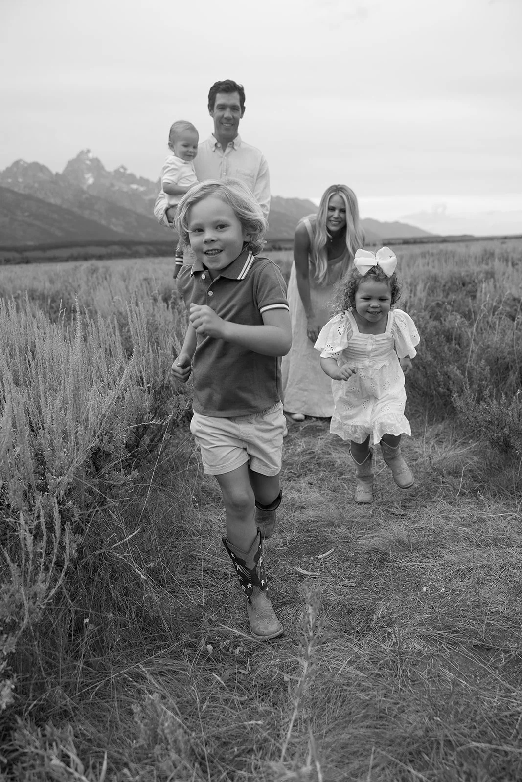 A family of five, including two children, a man, and two women, running with smiles in a field with mountains in the background.