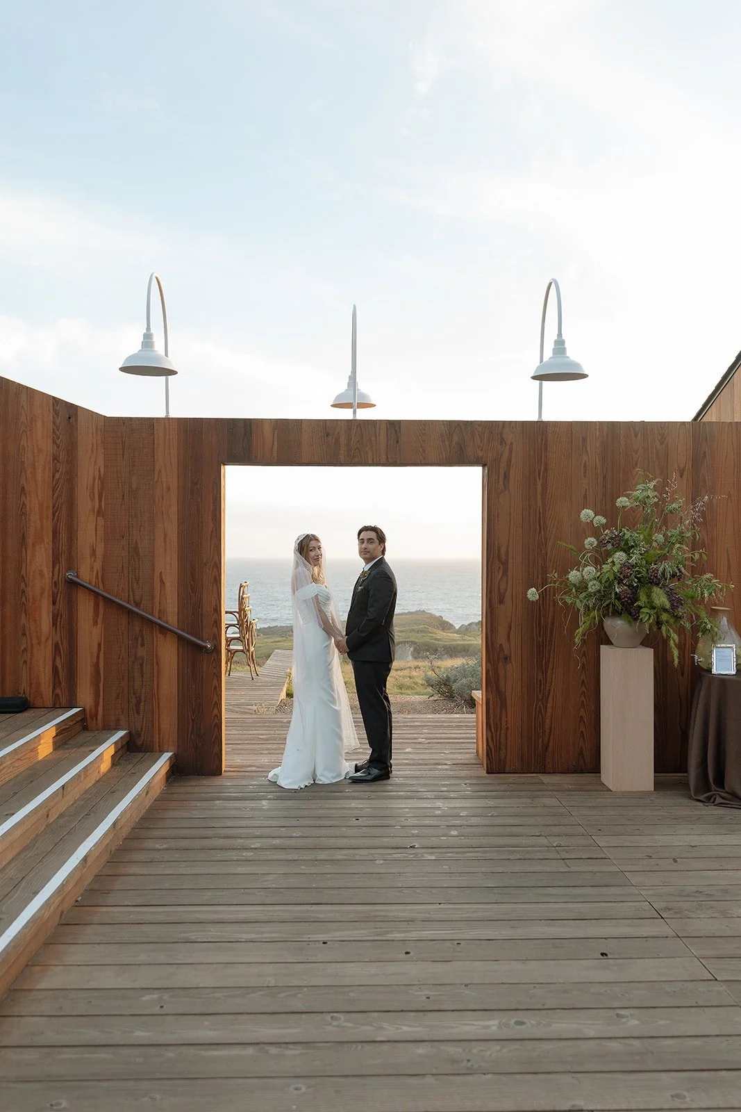 A bride and groom holding hands and facing each other on a wooden deck overlooking the ocean, framed by a wooden archway with decorative lighting and a large floral arrangement to the right.