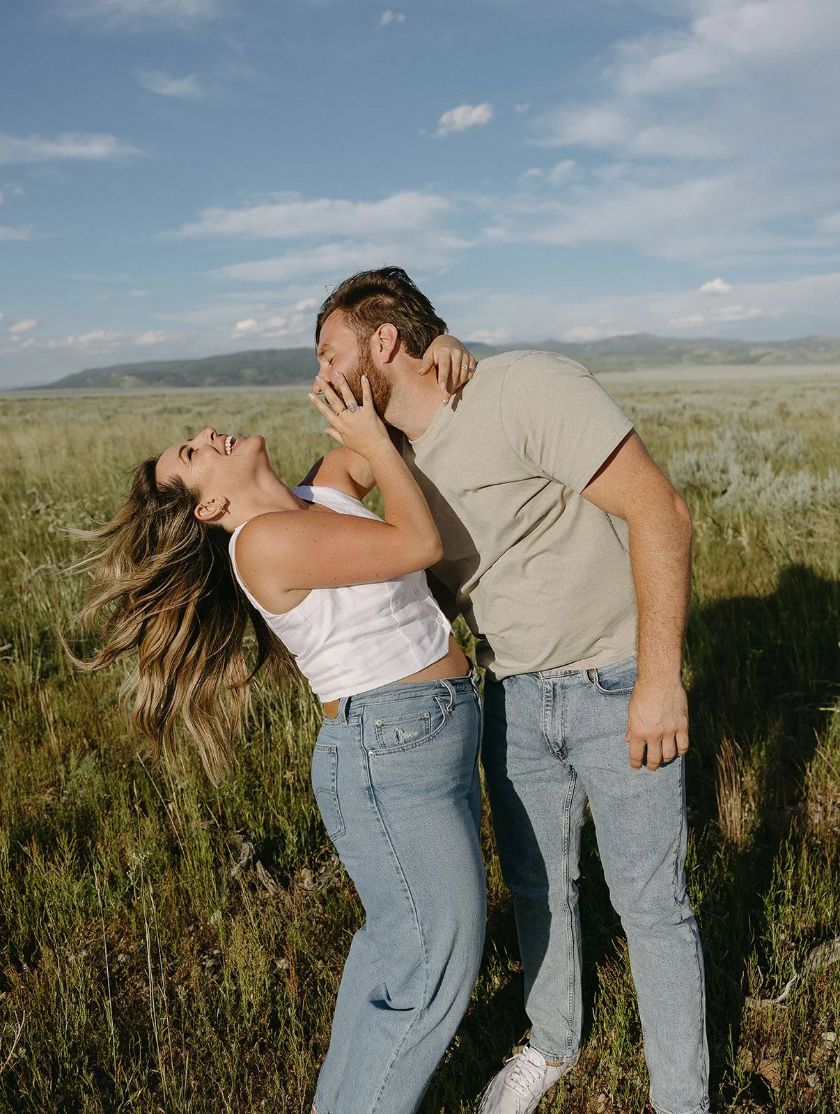 A couple is laughing and leaning in towards each other in a grassy field under a blue sky with clouds.