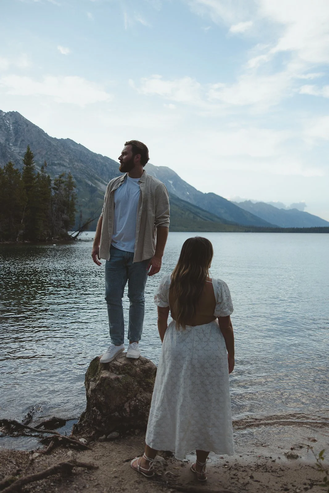 A man standing on a rock by a lake with mountains in the background, looking to the side. A woman stands at the water's edge with her back to the camera, wearing a white dress.