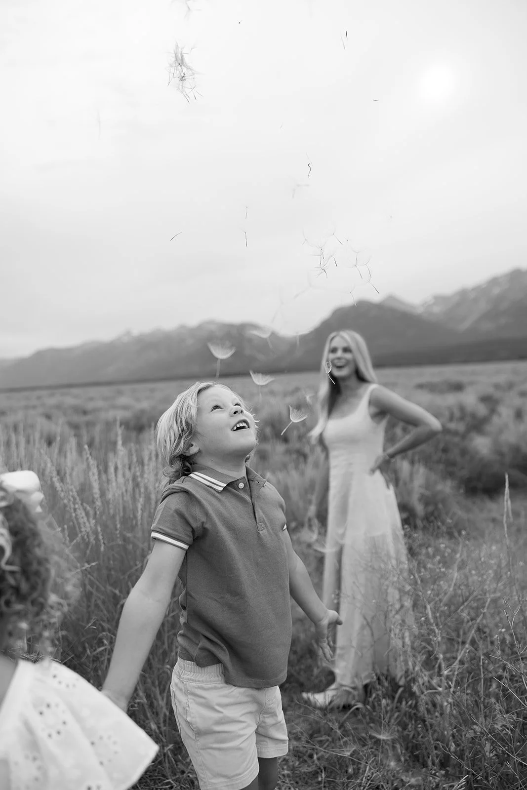 A young boy and a woman in a field of tall grass outdoors, with mountains in the background. The boy is looking up and smiling as he releases dandelions into the air, which are caught in a breeze. The woman stands behind him in a long dress, smiling 