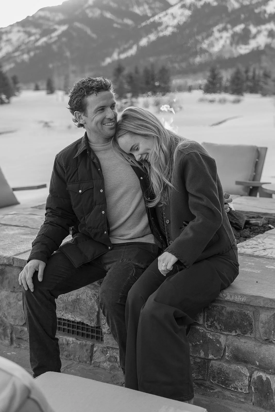 A man and woman sit close together outdoors in a snowy mountain landscape, smiling and laughing, with the woman leaning her head on the man's shoulder.