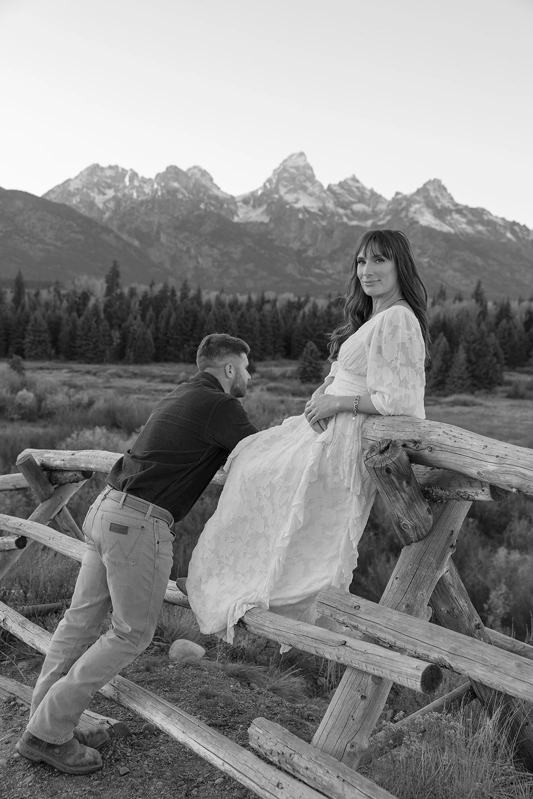 A woman in a white dress sitting on a wooden fence with a man kneeling beside her, in front of mountain scenery and trees.