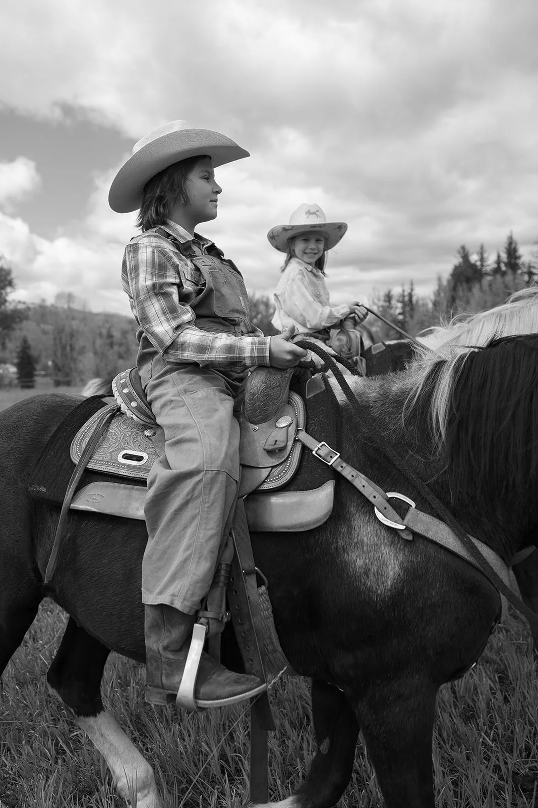 Two children wearing cowboy hats riding horses outdoors in a rural setting.