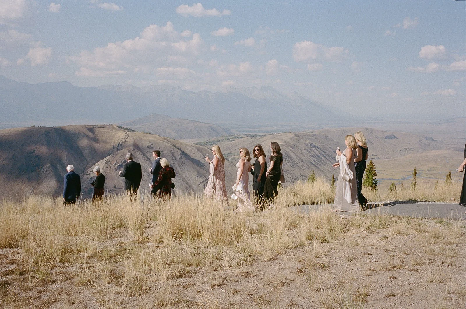 A group of people dressed in formal attire walking outdoors in a mountainous landscape with grass and hills under a partly cloudy sky.